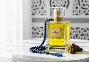 Botol parfum kaca kristal mewah dengan cairan berwarna emas, dikelilingi ornamen arabesque dan tasbih di atas meja marmer putih.