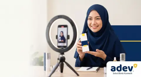 Foto profesional perempuan Indonesia berhijab tersenyum percaya diri sedang live shopping, memegang kemasan serum kosmetik premium ke arah kamera ponsel di atas tripod dengan ring light, latar meja kerja brand kosmetik rapi dan modern